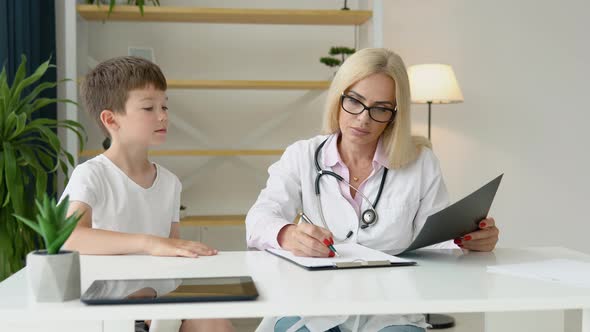 Smiling Senior Female Family Doctor in Uniform Consulting Little Kid Patient at Checkup Meeting in alt