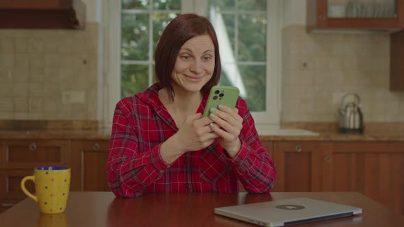 30s Woman in Red Shirt Reading Good News on Mobile Phone Sitting in the Kitchen alt