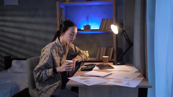 Young Asian Girl Student or Freelancer Working at Night at Home with Documents and Charts Drinking alt