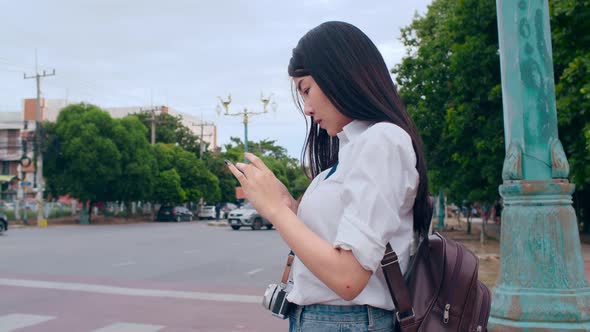 Asian backpacker woman tourist with camera stand in front of cross walk street look at mobile.