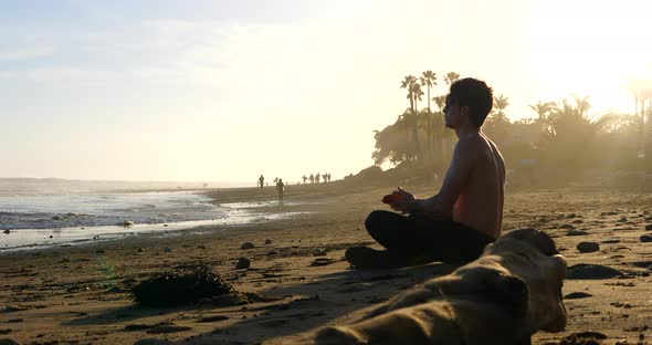 A muscular man watching the sunset in a meditation pose as surfers walk along the ocean water in sil alt