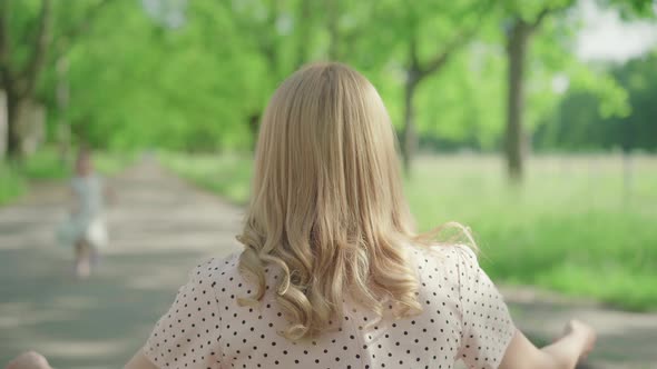 Back View of Blond Caucasian Woman Sitting on Alley in Summer Park As Children Running To Her alt