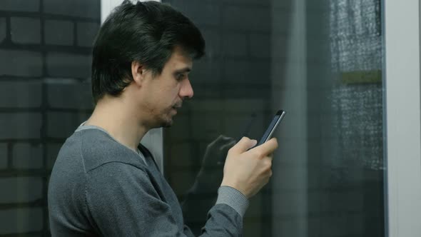 Man Is Browsing Internet Pages on Mobile Phone Standing Near the Window on the Balcony