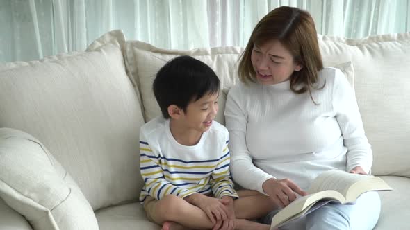 Asian Mother And Her Son Reading A Book On Sofa In Living Room Slow Motion  alt