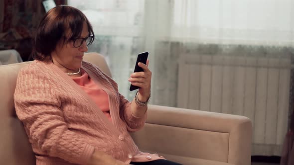 Grandmother In Glasses Scrolling On Screen On Cellphone. Mature Woman Holding Mobile Phone. alt