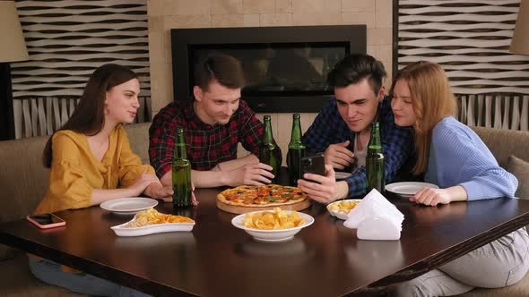 A Group of Friends Relax in a Cafe with Beer and Pizza They Look at the Phone alt
