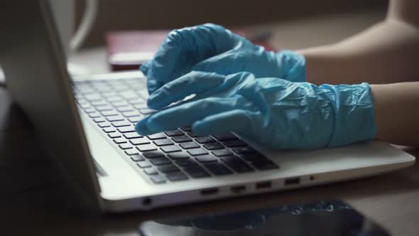 A Woman in Disposable Gloves and a Mask Works at Home with a Laptop alt