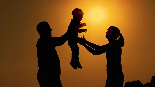Silhouettes at sunset.  Dad hands the little baby over to mom. mom rotates with the baby