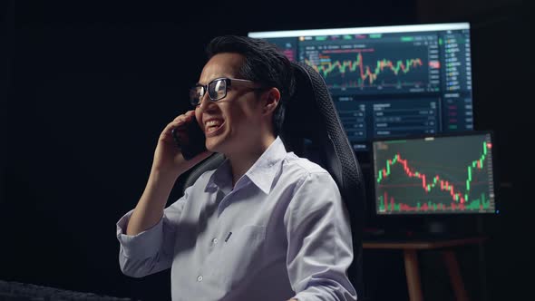 Asian Young Male Stock Market Broker Talking On Mobile Phone With Analyzing Graphs alt