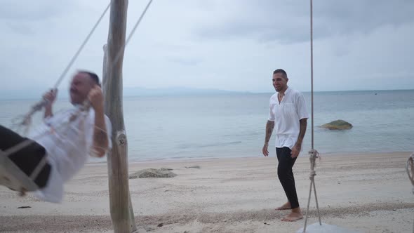Lovely Male Gay Couple Honeymooners Kissing While Riding a Beach Swing at Tropical Island at Dusk alt