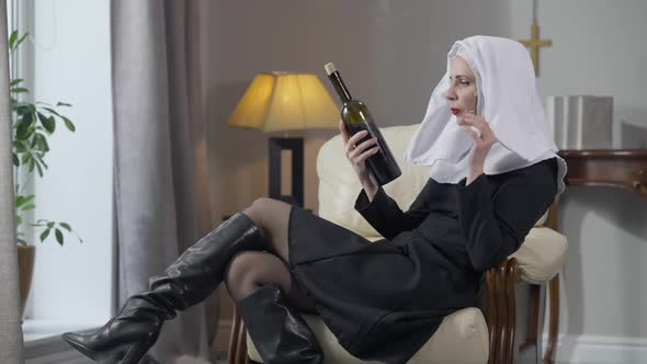 Side View of Concentrated Confident Woman in Nun Costume Sitting on Armchair Examining Wine Bottle alt