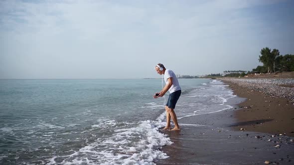 A Slender Whitehaired Elderly Man in White Headphones is Dancing Energetically Barefoot on the Beach