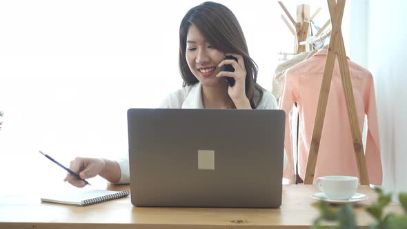 young smiling asian woman working on laptop while sitting in a living room using phone. alt
