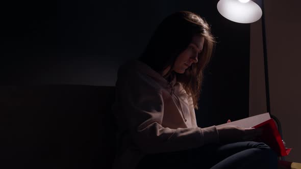 Attractive Longhaired Girl Reading Book Sits In Dark Room at Home on Sofa