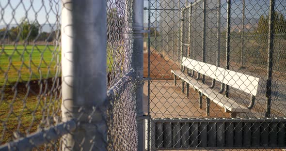 Slow pan across a chain link fence and an empty baseball field bench in the dugout of a local park a alt