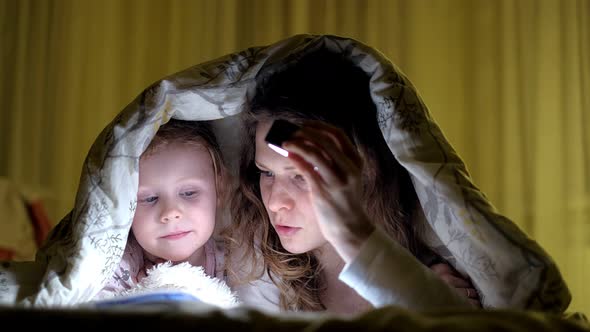 Smiling Mother and Her Daughter Reading Book Under Bed Cover and Holding a Flashlight