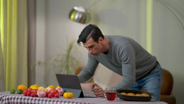 Young Focused Middle Eastern Man Watching Online Recipe on Tablet Standing at Table with Fruits and alt