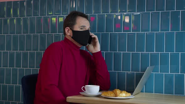 Middleaged Man in Black Protective Mask with Laptop in Cafe Talking on Mobile