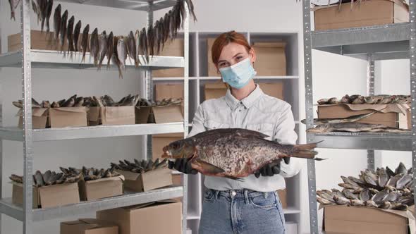 Portrait of Young Woman in Medical Mask with Large Sea Fish in Her Hands Background of Shelves with alt