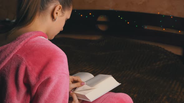 Young Woman in Pink Pajamas Sitting Reads a Book alt