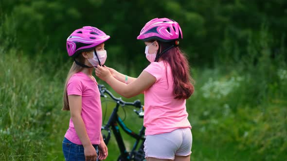 Two little girls in pink helmets and medical masks. The sister helps the younger girl put on her 