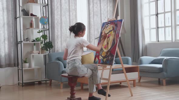 Asian Artist Girl Sitting And Holding Paintbrush Painting A Girl's Hair On The Canvas alt