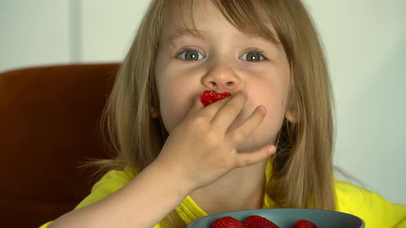 Little Girl Sits By Table and Eats with Appetite Ripe Strawberry on Kitchen