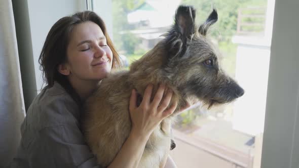 Woman with Dark Hair Hugs Grey Dog Looking Outside Window