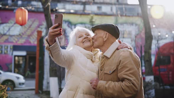 An Elderly Couple Takes Selfies in Which They Kiss While Standing on the Street