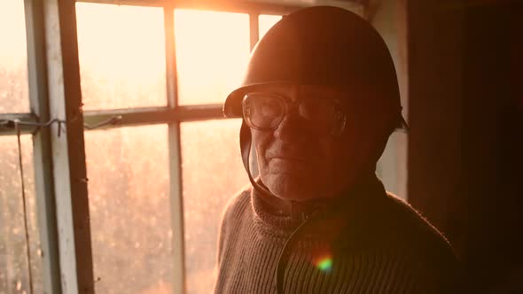 Elderly Man with Glasses Picks Up Military Helmet in Dust and Putting Helmet on Head in House Window alt