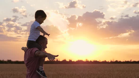 A Happy Father Plays with His Son While Running Through a Wheat Field. A Small Boy Sits on His alt