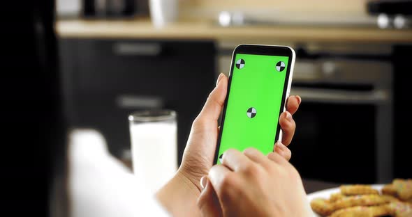 Woman Holds a Smartphone with a Green Screen in Her Hands Sitting in the Kitchen