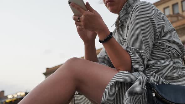 A Woman in a Striped Dress and with Beautiful Bare Legs Sits Outdoors and Holds a Smartphone in Her