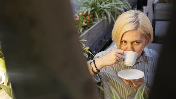 An Adult Young Caucasian Blonde Drinks Coffee From a White Cup in a Summer Restaurant at a Table in