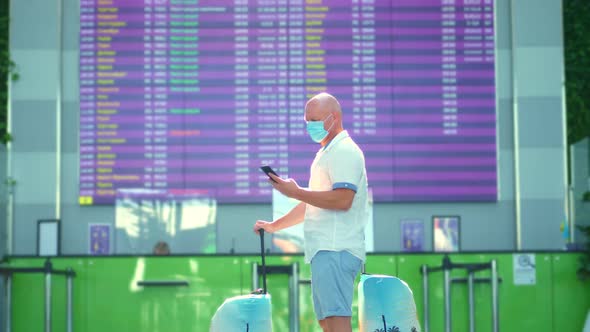 Male Air Passenger in Medical Mask, with Luggage, Standing Against Departure Board at Airport alt