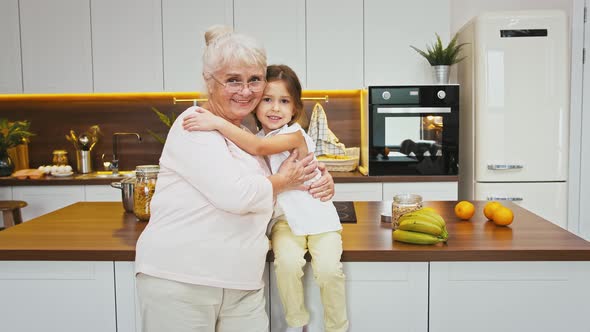 Grayhaired Granny and Little Granddaughter are Smiling and Hugging While Posing Against a Big Table alt