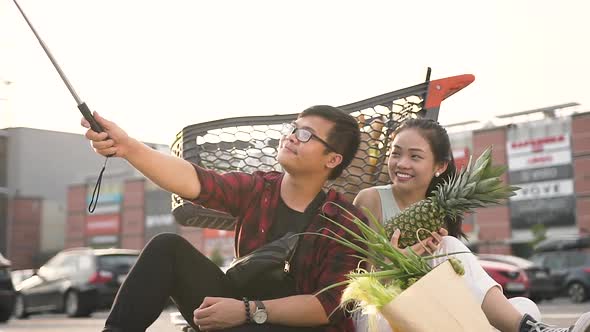 Happy Vietnamese Couple Sitting on the Ground Near Big Shopping Centre and Making Selfie alt