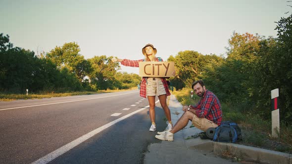Couple of Travellers Hitchhiking on Empty Road Man Resting on Ground alt