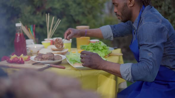 Side View of Handsome Concentrated African American Man Putting Hot Grilled Patties on Burgers on alt