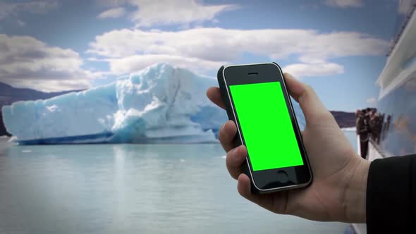 Hand holding an Old Smartphone with Green Screen near an Iceberg. alt
