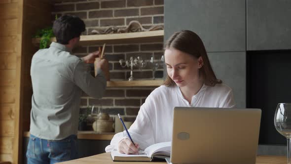 Young Woman Using a Laptop in the Kitchen and a Man Prepares Food alt