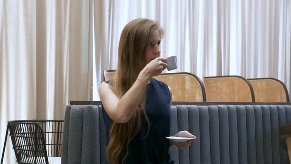 Brooding Young Woman Drinking Coffee From Cup Sitting on the Sofa in Cafe alt