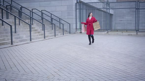 Young Happy Caucasian Woman in Red Coat and High Black Boots Walking Along the City Square and alt
