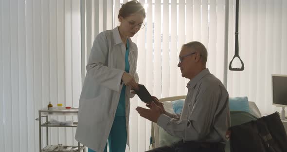 Aged Man Patient Sitting on Bed in Hospital Ward and Signing Discharge Form alt
