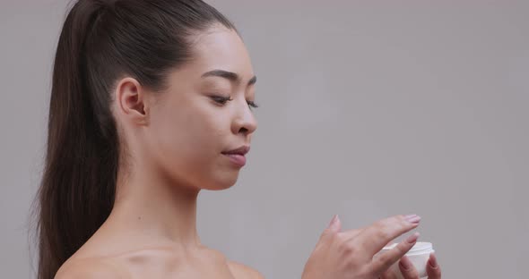 Young Asian Woman Opening Jar and Applying Moisturizing Cream on Face alt