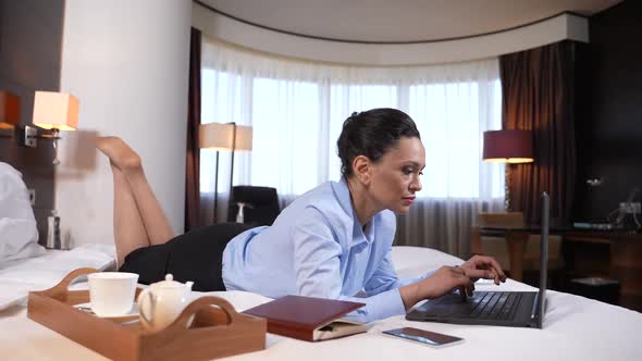 Woman Networking on Laptop Lying on Bed in Suite alt