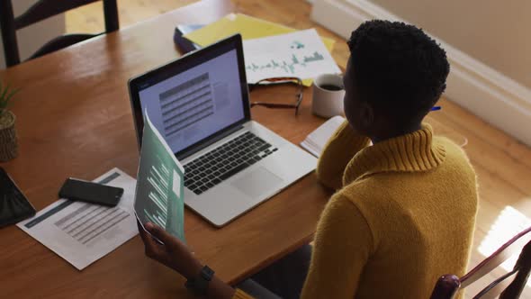 Overhead view of african american woman reading document and using laptop while working from home alt