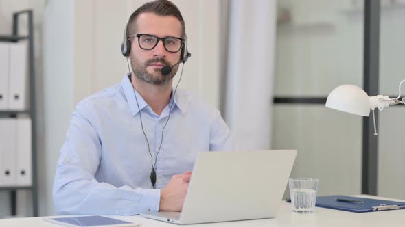Middle Aged Man with Headset Looking at Camera While Using Laptop alt