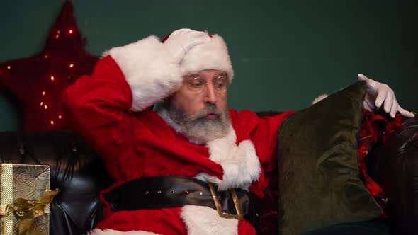 Tired Santa Claus Sleeps on the Couch Wakes Up Checks If Gifts are on the Spot and Falls Asleep alt