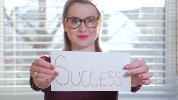 A Young Blonde Female With Glasses Holding Up A Piece Of Paper That Reads Success 1